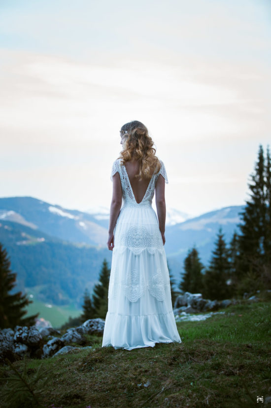 Braut blickt in die Bergwelt des Wildenkaiser, Abendstimmung, weiches Licht, Bohokleid, tiefer Rücken, blonde lange Locken, Portfolio, Vintage-Bohème, Brautkleider ,Styled Shoot, Wilder Kaiser, Crème Bohème, Hochzeitskleider, Ernst Merkhofer, Fotograf, Brautkleid Inspiration , Brautkleid, Spitze, blue hour, Natur, Berge, Hochzeiten