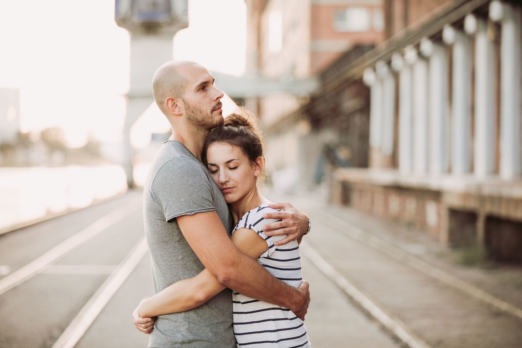 junges Paar steht engumschlungen in einer Industrielandschaft, Kai oder Hafen. Die junge Frau hat Ihren Kopf an die Brust des Mannes gelegt und die Augen geschlossen. Portfolio, Portraits