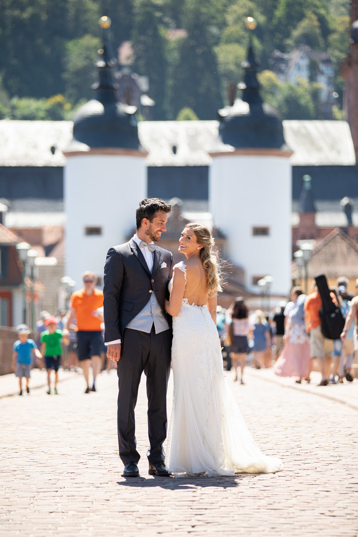 Kleiner Spaziergang zu den Highlighs der Heidelberger Altstadt bei ihrer After Wedding Session. Brautpaar steht mitten auf der Alten Brücke in Heidelberg, Touristen im Hintergrund. Die Zwillingstürme des Brückentors erstrahlen frisch renoviert. Braut ist dem Bräutigam zugedreht und schaut über die Schulter in die Kamera. Der Bräutigam blickt sie an. .