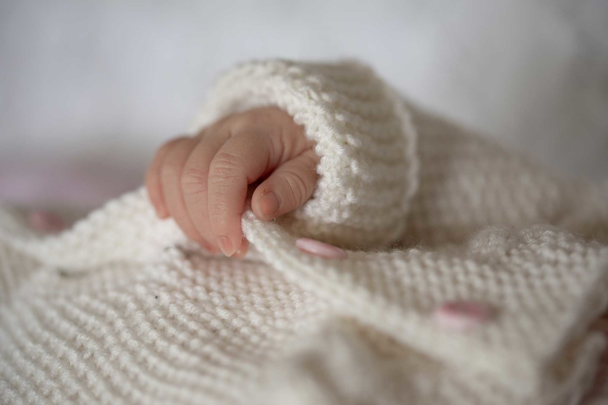 Babyhändchen schaut aus dem Ärmel einer weißen Strickjacke heraus, das Baby liegt auf dem Rücken und die Hand und der Arm ruhen auf dem Bauch. Die kleinen Finger halten die Knopfleiste mit den rosafarbenen Knöpfen ganz leicht fest. Portfolio
