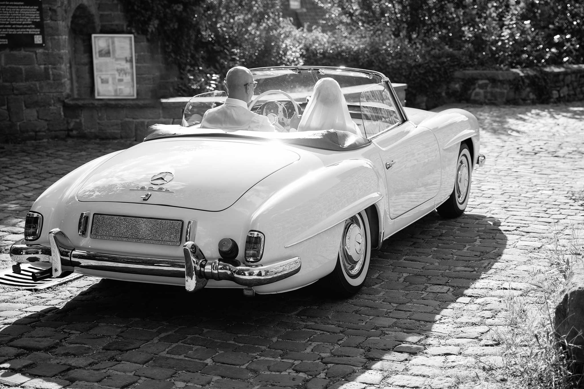 frisch vermähltes Brautpar fährt im weißen Oldtimer Cabrio auf einem Kopfsteinpflaster im Gegenlicht in eine neue Zukunft. Hochzeiten, Portfolio, Hochzeitsauto, 190SL Mercedes-Benz